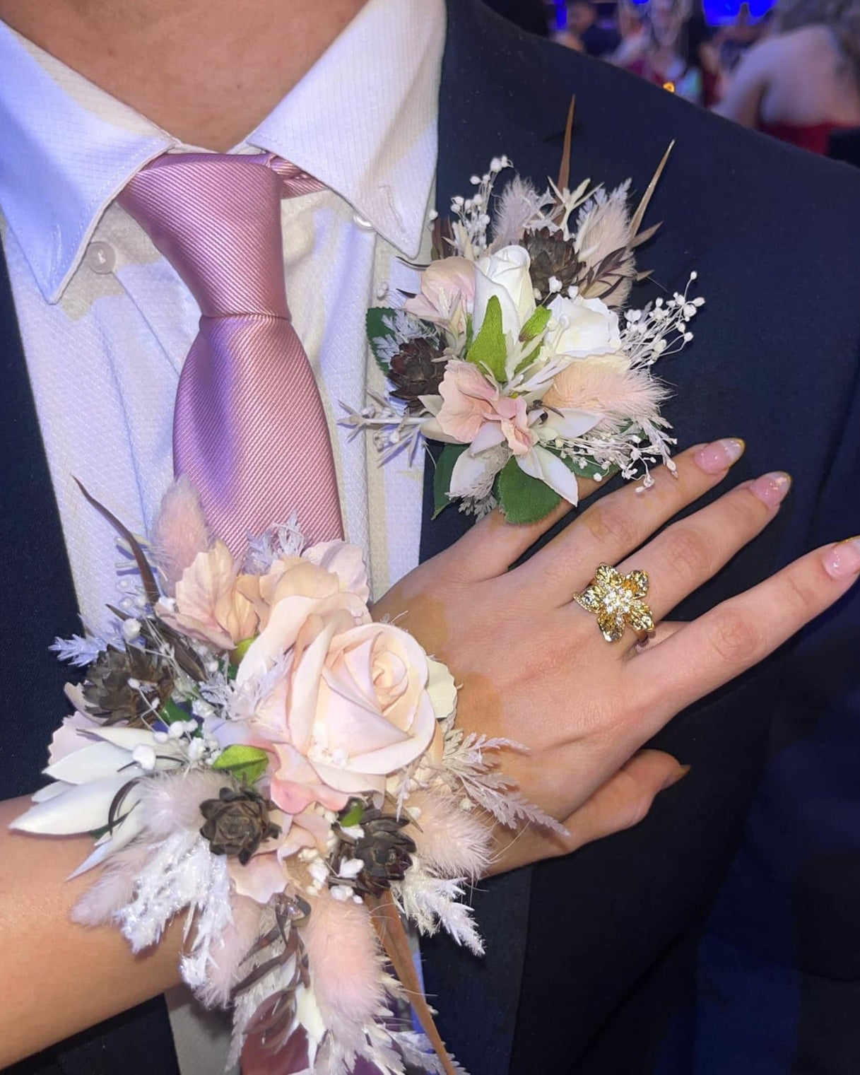 Person wearing a formal outfit with a floral corsage and ring, blurred background